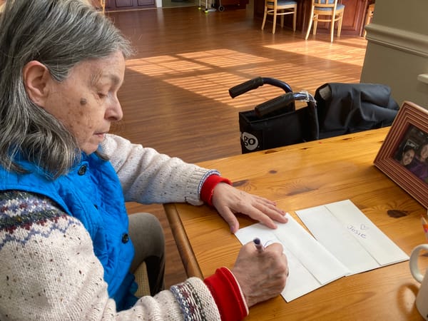 A woman in a sweater and blue vest writes at a wooden table.